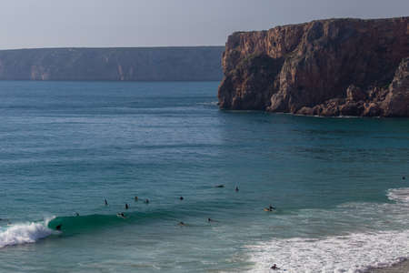A landscape of sea with people surfing on the waves surrounded by rocks under a blue sky in Portugalの写真素材