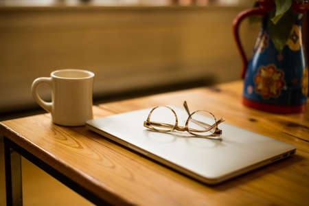 A wooden desk with a laptop a mug and glasses in a working environmentの写真素材