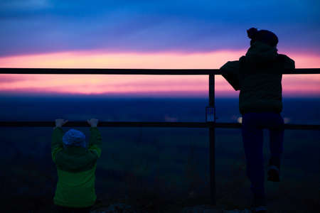 A picture of two kids watching the beautiful sunset near the fences surrounded by greeneryの写真素材
