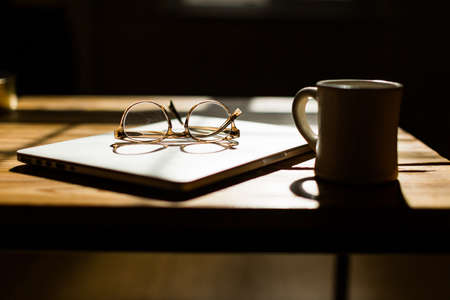 A wooden desk with a laptop a mug and glasses in a working environmentの写真素材