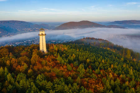 An aerial shot of a white lighthouse in the middle of a tree forest near high grassy hills covered...