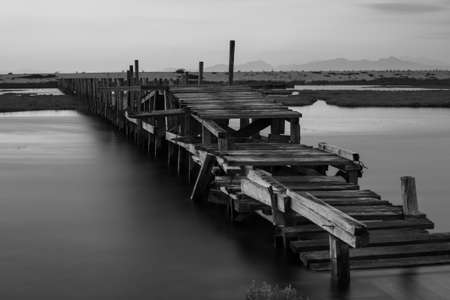 A grayscale shot of a wooden dock on the calm sea with high mountains in the backgroundの写真素材