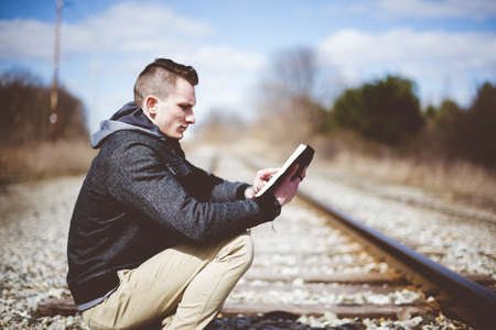 A shallow focus shot of a male sitting down on train tracks while reading the bible