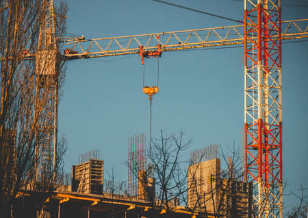 A white and red construction crane surrounded by buildings and greenery under a blue sky and...