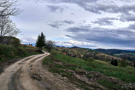 A road surrounded by hills covered in forests with houses under a cloudy sky on the background