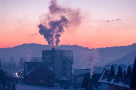 A smoke and houses covered in the snow with mountains and forests on the background during the sunsetの写真素材