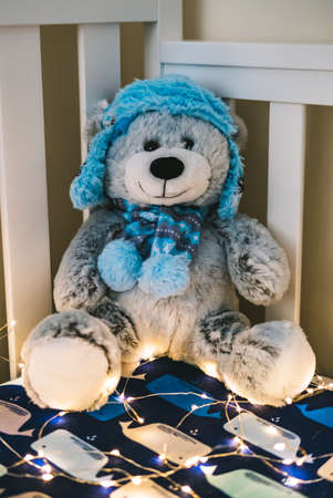 A grey and blue teddy bear on a white bed covered with Christmas lightsの写真素材