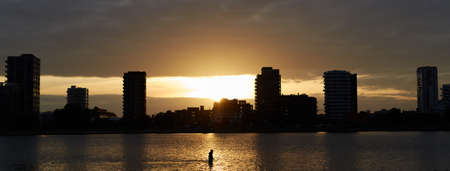 A landscape of a river with a person in it surrounded by modern buildings during sunset