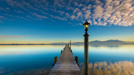 A wooden bridge and a lamp in a sea with mountains on the background under a cloudy sky during sunsetの写真素材