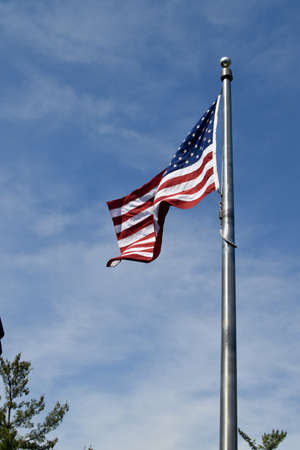 A low angle view of the American flag near trees under a blue cloudy sky and sunlight