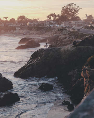 A vertical shot of a person standing on the cliff near the sea with buildings in the distanceの写真素材