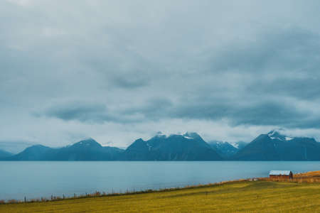 A field covered in greenery surrounded by the sea and mountains covered in the snow under the cloudsの写真素材