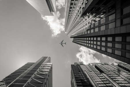 A low angle greyscale shot of an airplane flying above high rise buildingsの写真素材