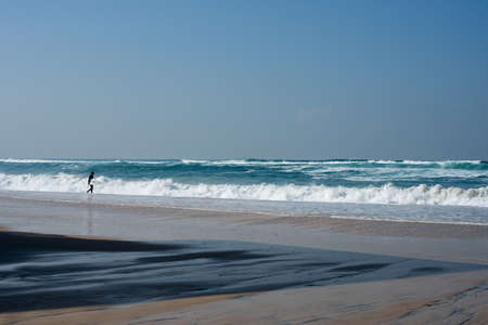 A beach surrounded by the wavy sea with a surfer near it under a blue sky and sunlightの写真素材