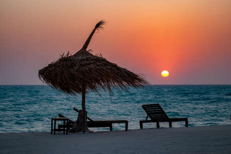 A folding chair and a weaved parasol on the beach with the beautiful view of the sunset in the backgroundの写真素材