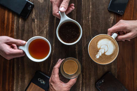 A high angle shot of four phones on the table and people holding cups with different types of coffeesの写真素材