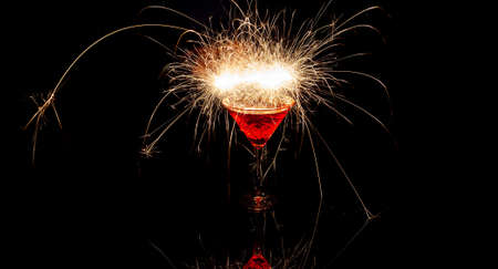A glass cup of red cocktail with sparks reflecting in glass with a black background