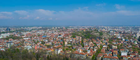 A high angle view of a city covered in greenery and buildings with red roofs under a blue skyの写真素材