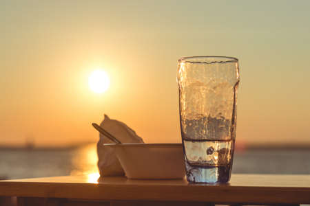 A glass cup filled with water and a bowl on a table with the beautiful view of sunset in the backgroundの写真素材