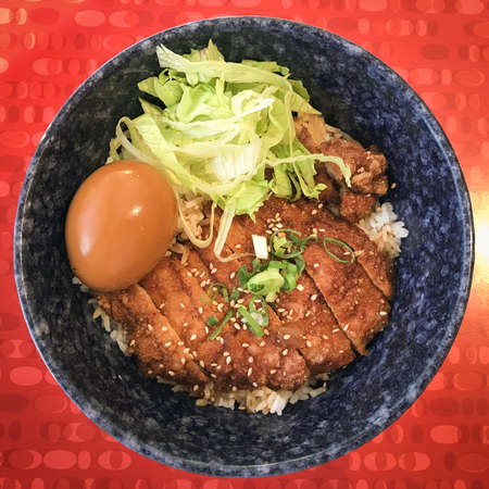 A high angle shot of fried meat with rice, cabbage and an egg in a bowl on a red surfaceの写真素材