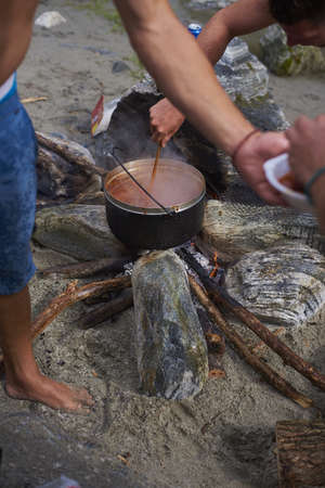 A closeup look of people making a dish in a black metallic pot on a bonfire surrounded by sand