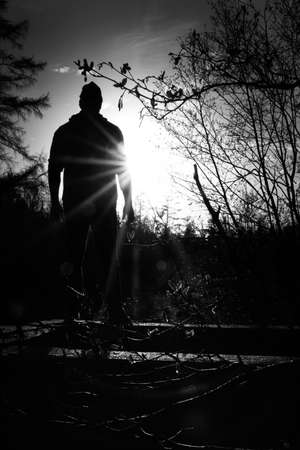 A vertical grayscale shot of a male standing near leafless trees with the sun shining in the backgroundの写真素材