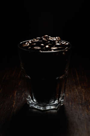A vertical shot of a glass cup filled with coffee beans on a wooden surface with a black backgroundの写真素材