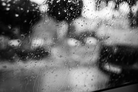A greyscale shot of a glass covered with raindrops with a blurred background
