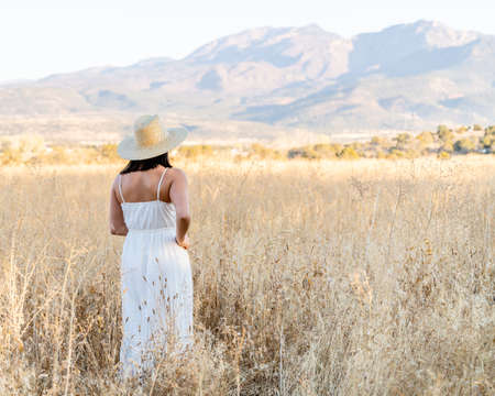 A beautiful view of a woman in a white dress and a hat standing in a wheat field with mountains in the distanceの写真素材