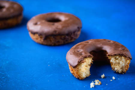 A closeup shot of chocolate-dipped half-eaten doughnut on a blue surface with blurry doughnuts in the backgroundの写真素材