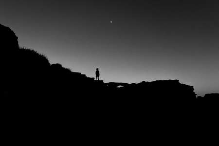 A grey scale shot of the silhouette of a person standing on big rocks