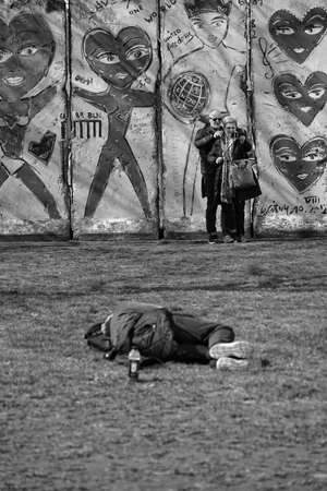 An older couple prepares for a selfie in a park. Behind them a wall sprayed with graffiti reads: "United freedom". a drunken man lies in front of themのeditorial素材