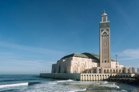 The Hassan II Mosque surrounded by water and buildings under a blue sky and sunlightのeditorial素材