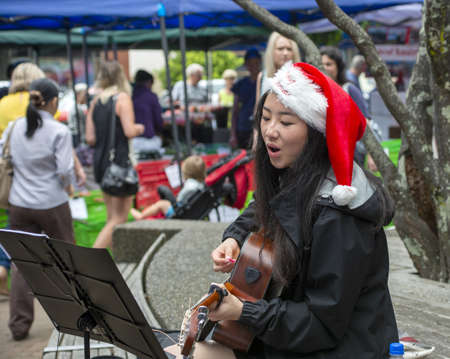 AUCKLAND, NEW ZEALAND - Dec 12, 2015: Auckland, New Zealand, december 12 2015. An attractive young asian woman wearing a christmas hat and playing a guitar and busking at a farmers marketのeditorial素材