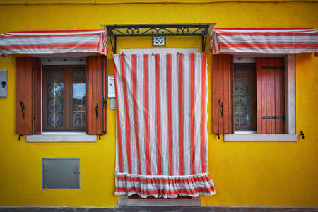BURANO, ITALY - Apr 29, 2017: Horizontal shot of a colorful house with yellow walls and white and orange striped curtains in front of the entrance door in Burano, Italyのeditorial素材