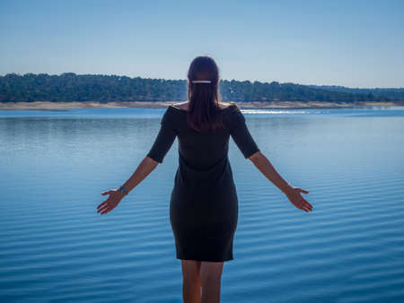 A young female in a nice black dress standing with her arms open enjoying the beautiful morning view of the seaの写真素材