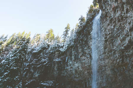 A waterfall surrounded by rocks and evergreens covered in the snow under the sunlightの写真素材