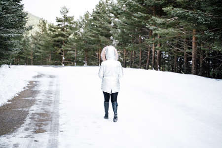 A wide shot of a woman walking towards a path on a snowy road surrounded by treesの写真素材