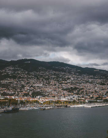 A vertical shot of buildings on the mountain under a cloudy sky in Funchal, Madeira, Portugal.の写真素材