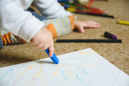 A closeup shot of a baby drawing with a blue crayon sitting on the floorの写真素材