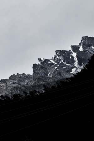 A vertical shot of the silhouettes of trees and cable lines under the snowy rocksの写真素材
