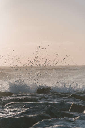 A vertical shot of sea waves hitting the rocks under a cloudy sky