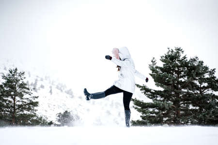 A wide shot of a woman kicking snow and having fun on a snowy hill with trees around on a winter dayの写真素材