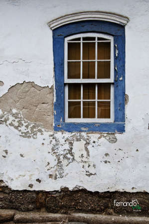 A vertical shot of a white old and cracked wall with a single blue window on it in Paraty Brazilの写真素材