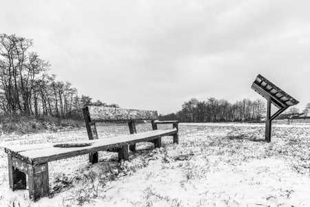 A grayscale shot of benches on a field covered in snow under a cloudy skyの写真素材