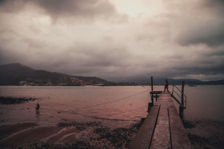 A wooden bridge on a beach surrounded by the sea and hills covered in greenery and fog