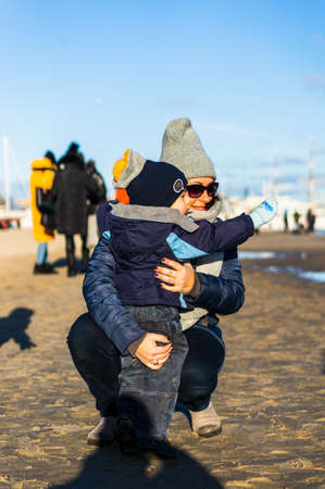 GDYNIA, POLAND - Jan 11, 2020: Adult woman with sunglasses wearing warm clothes kneeling next to a small boy pointing at something on a beach on a cold winter day.の写真素材