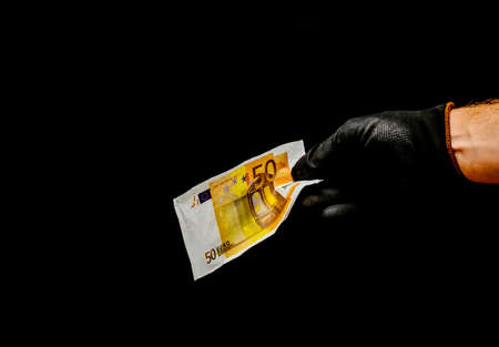 A landscape shot of a man wearing black gloves holding a 50 euro note with a pitch-black background
