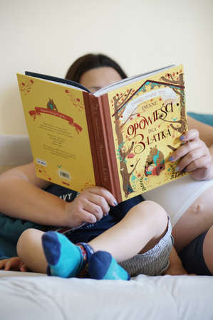 POZNAN, POLAND - Jul 17, 2017: Pregnant woman reads from a fairy tales book to a child on a sofaのeditorial素材