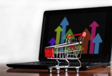 A closeup shot of a small shopping cart with a laptop in the background - internet commerceの写真素材